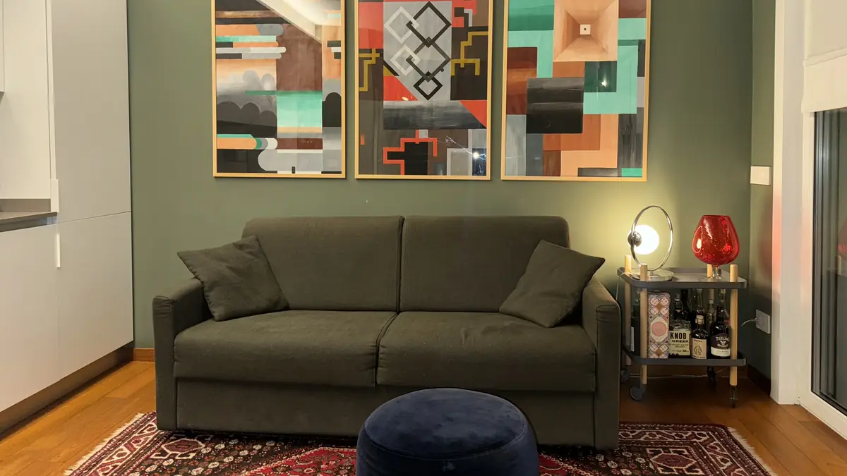 The living room with sage-green walls, the sofa, a red rug and three colourful paintings